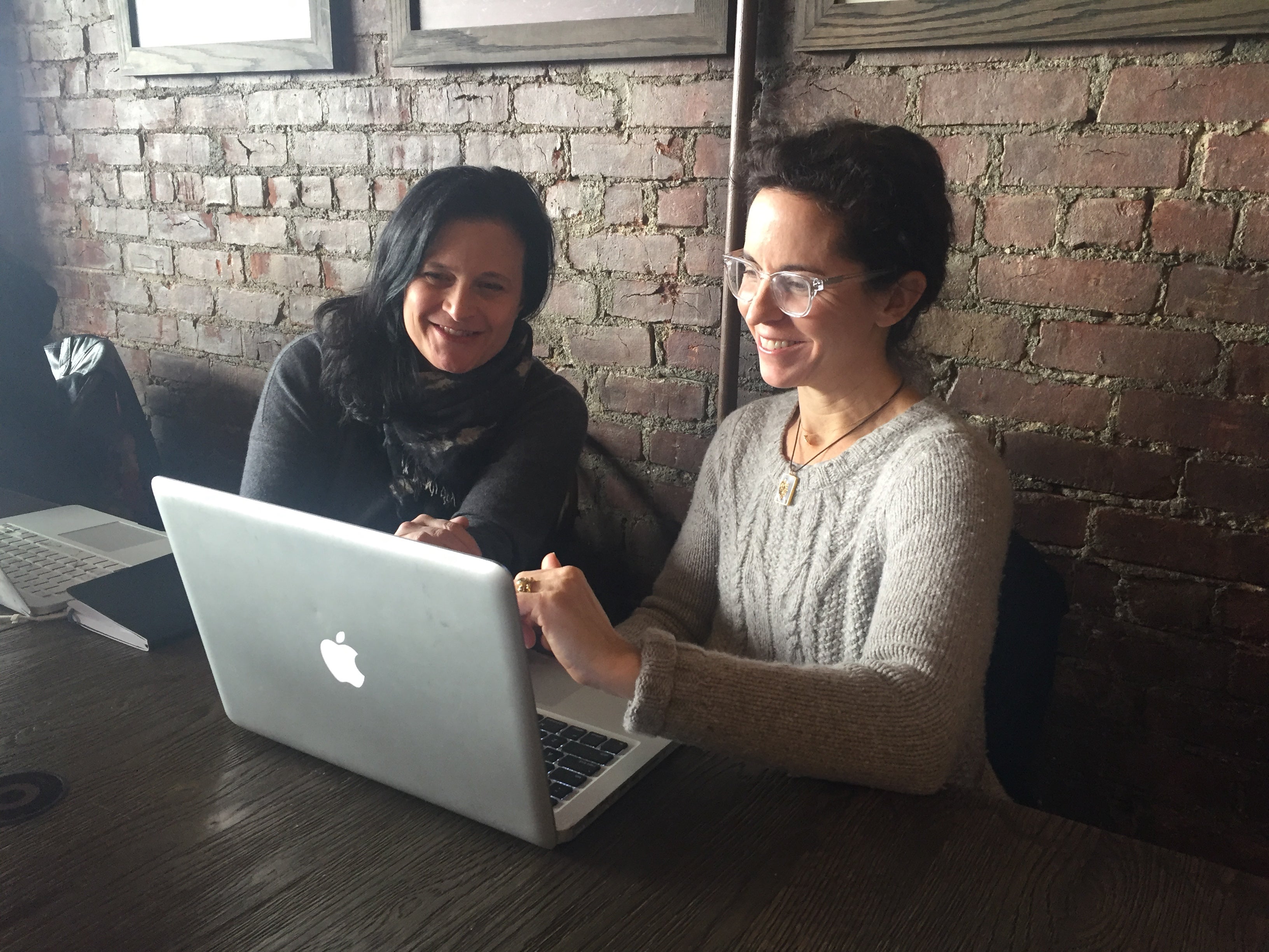 Two Women Smiling and working on laptops in front of brick wall in coffee shop