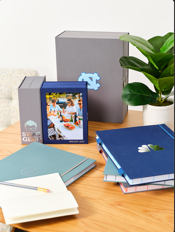 Savor keepsake boxes  and fan folios on top of a table with a notebook; the fan folios are personalized with logos